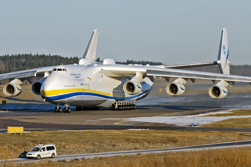 A photo of the world's largest plane, the Antonov AN-225. It is sitting in an airfield. The plane is mostly white and has yellow and blue stripes.