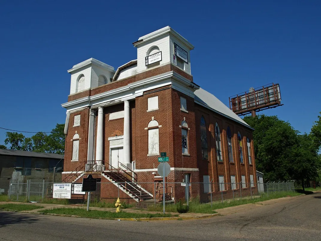 A stately red brick and white trim building, run down and surrounded by chain link fence, on the corner of a city block