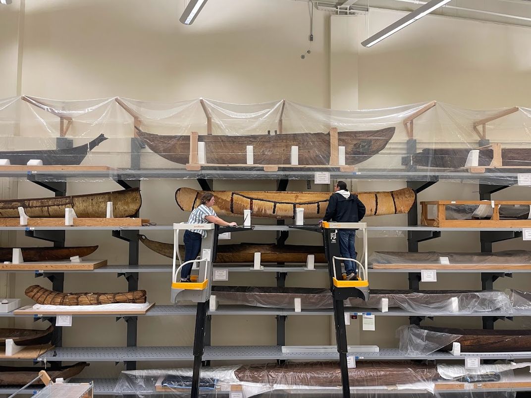 Two people on lifts in front of a shelved wall of canoes and other watercrafts.
