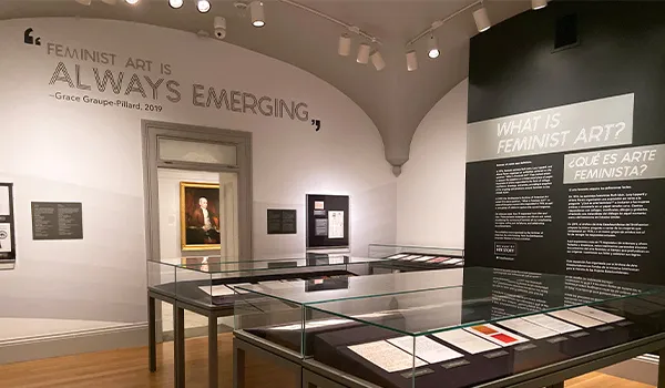 An image of the 'What is Feminist Art' exhibition with wall quote reading 'Feminist art is always emerging' and a portrait of a man seen through the doorway