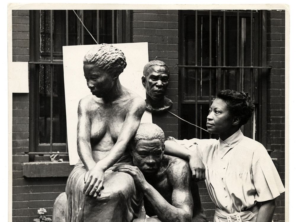A black and white photograph of artist Augusta Savage standing next to her sculpture