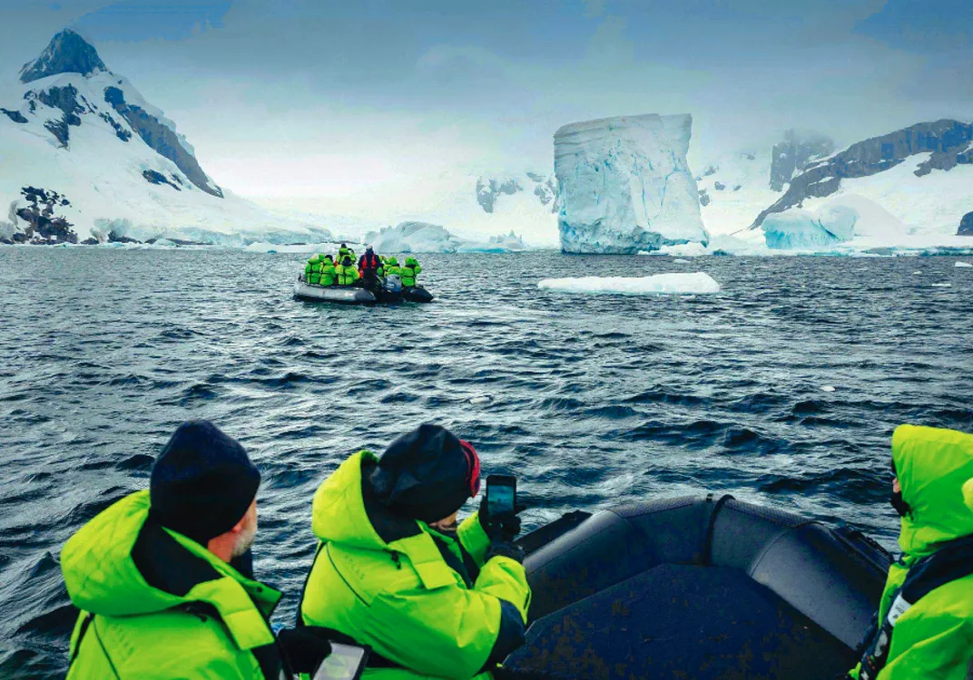 Tourists wearing bright green jackets are on two inflatable boats in icy waters, observing a large iceberg and snow-covered mountains.