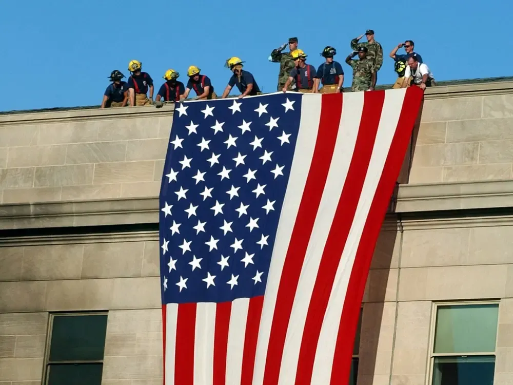 Several men stand on the roof of a large building, unfolding a red, white, and blue flag with a stars-and-stripes pattern over the side of the building.