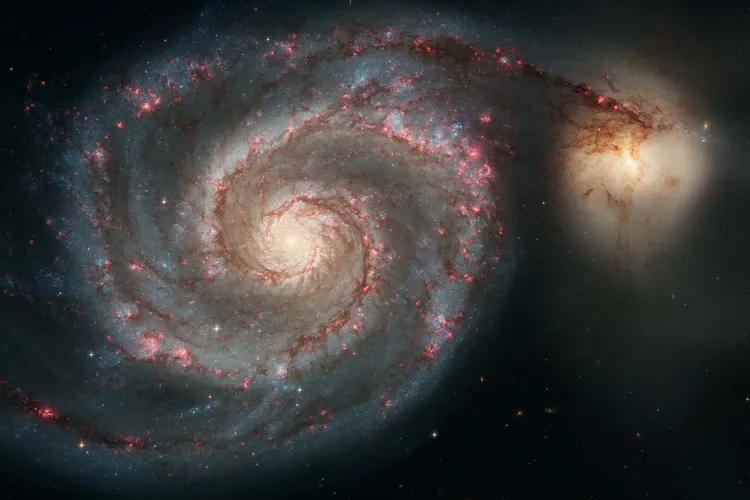 a swirling spiral galaxy with a yellow center and arms with blue and pink, one arm stretches and interacts with another galaxy