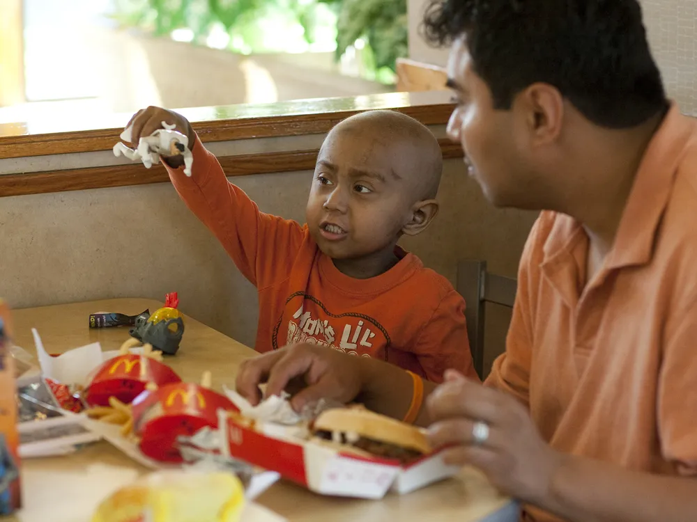An image of a 5-year old child playing with a McDonald's Happy Meal toy. In front of the child is a happy meal with fries and seated next to the child is his father.