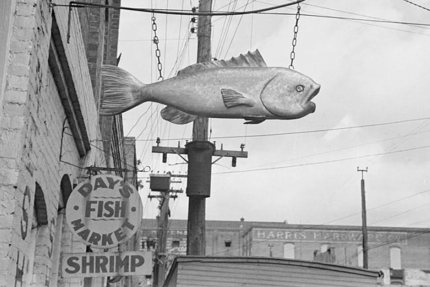 Along a brick storefront hangs a sign carved or painted to look like a fish with its mouth gaping. Beyond it, a sign with text Day's Fish Market, Shrimp. Black-and-white photo.