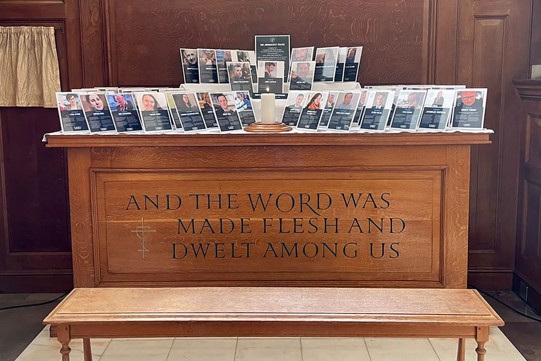 A wooden altar containing a single lit candle and several small memorial cards, each with a profile photo and text. Text inscribed on the side of the altar: AND THE WORD WAS MADE FLESH AND DWELT AMONG US.