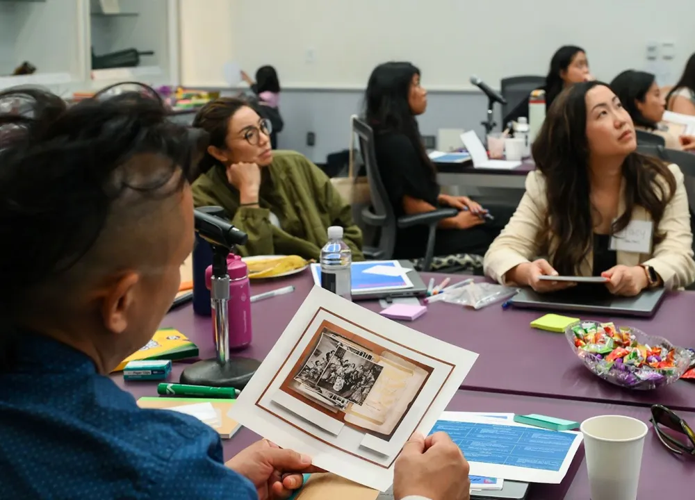 A man in a blue shirt looks at a photograph of archival images while several other people look toward a presentation