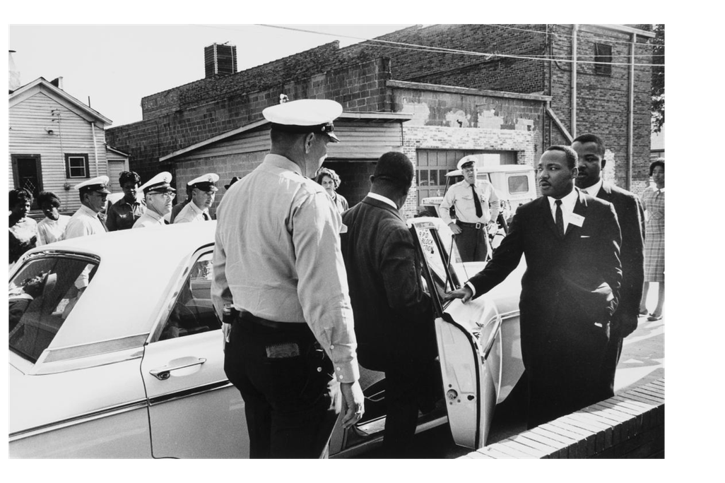 A black and white photograph of policeman in foreground, one man stepping into a squad car and MLK with an arm outstretched moving toward the car