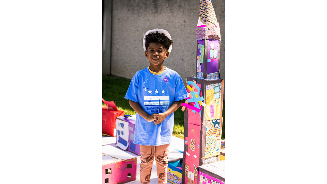 A student wearing a blue DCPS t-shirt and hardhat stands with his hands clasped. He is smiling and standing next to his cardboard skyscraper, which is a foot taller than him.
