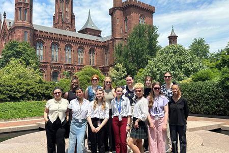 A group of thirteen people stand together in front of green shrubs and the Smithsonian Castle.