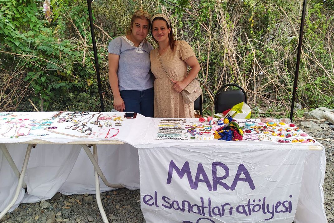 Two women pose behind a table with jewelry and small crafts for sale. A hand-painted sign on the front reads MARA, with words in Turkish.