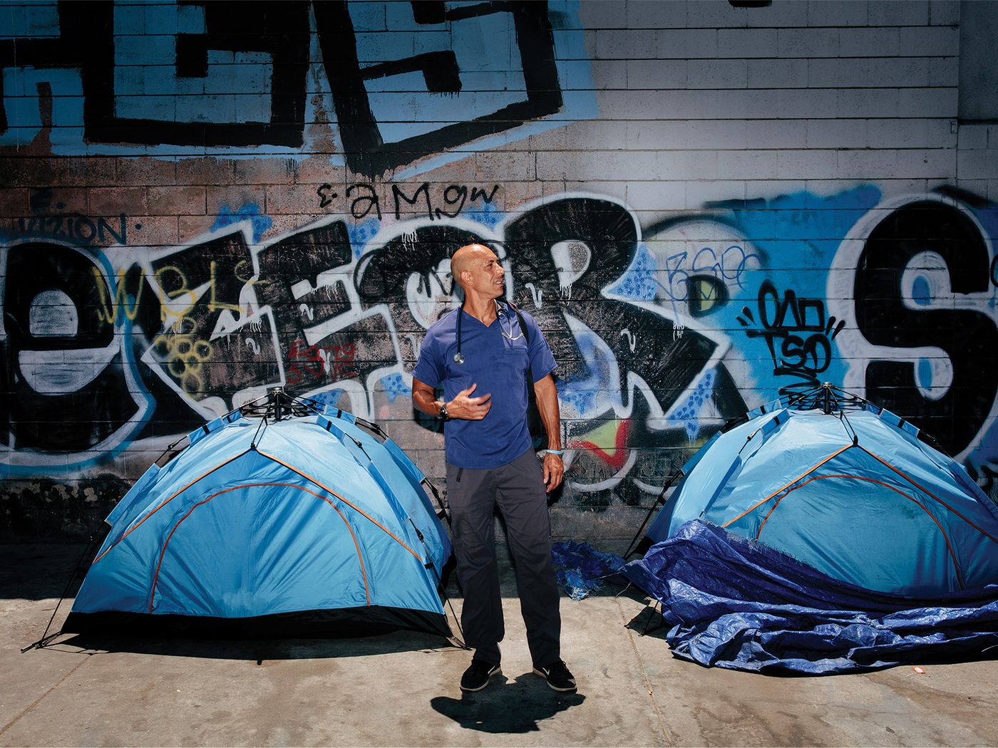 man standing in front of two blue tents and a wall of graffiti