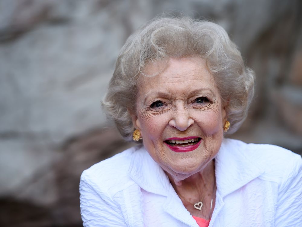 White, a white woman with curly white hair, smiles at the camera in a bright white suit with a heart shaped necklace