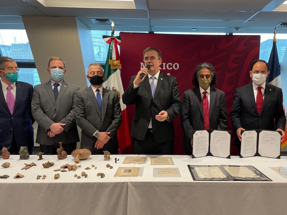 Seven people in suits and ties stand in front of a table with antiquities and 16th century documents laid out