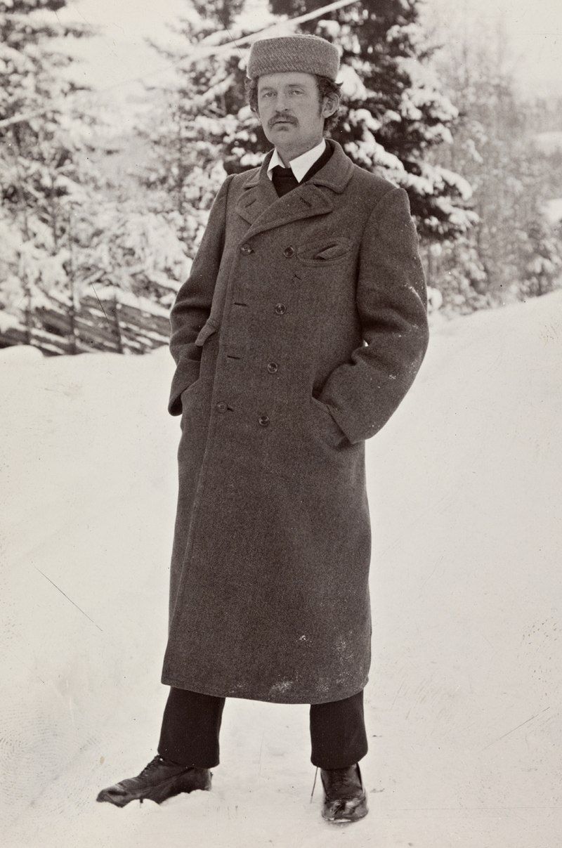 The artist, a white man with a mustache, in a long winter coat standing in a field covered in white snow