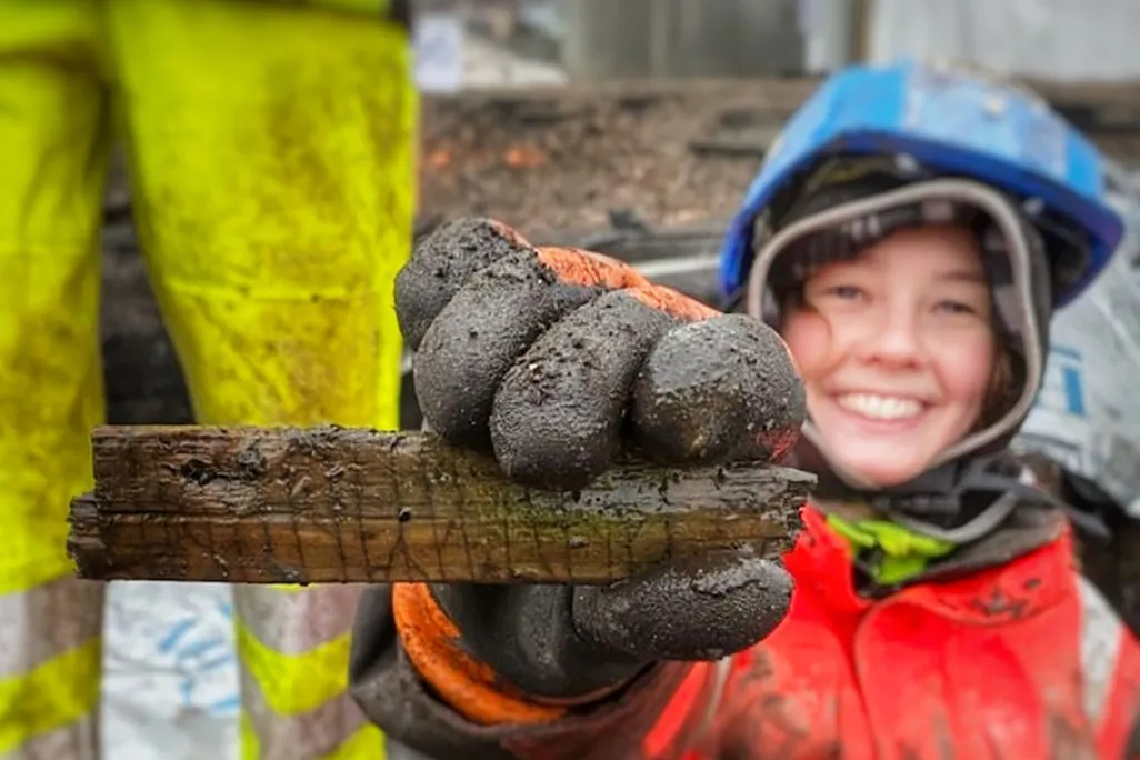 A white woman smiles and holds out the wooden slat, which is about the length of her hand and bears angular markings