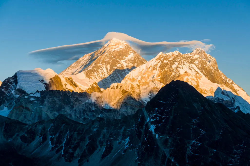 A landscape photograph of Mount Everest. The bottom half of the mountain is in the shade, but the sun warms the top half. Its peaks and valleys are covered with snow, and gray rocks emerge from beneath. The sky is bright blue and has a small white cloud.