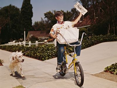 A boy riding his bike while delivering newspapers with his dog in tow, 1970s