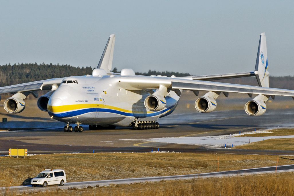 A photo of the world's largest plane, the Antonov AN-225. It is sitting in an airfield. The plane is mostly white and has yellow and blue stripes.