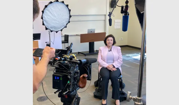 Annamarie Lusardi, seated in a chair facing a camera and lighting equipment.