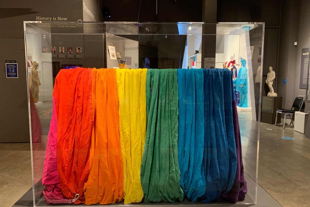 A view of a museum display case with the gathered folds of a piece of rainbow-striped cloth, including 8 bright colors from pink to purple