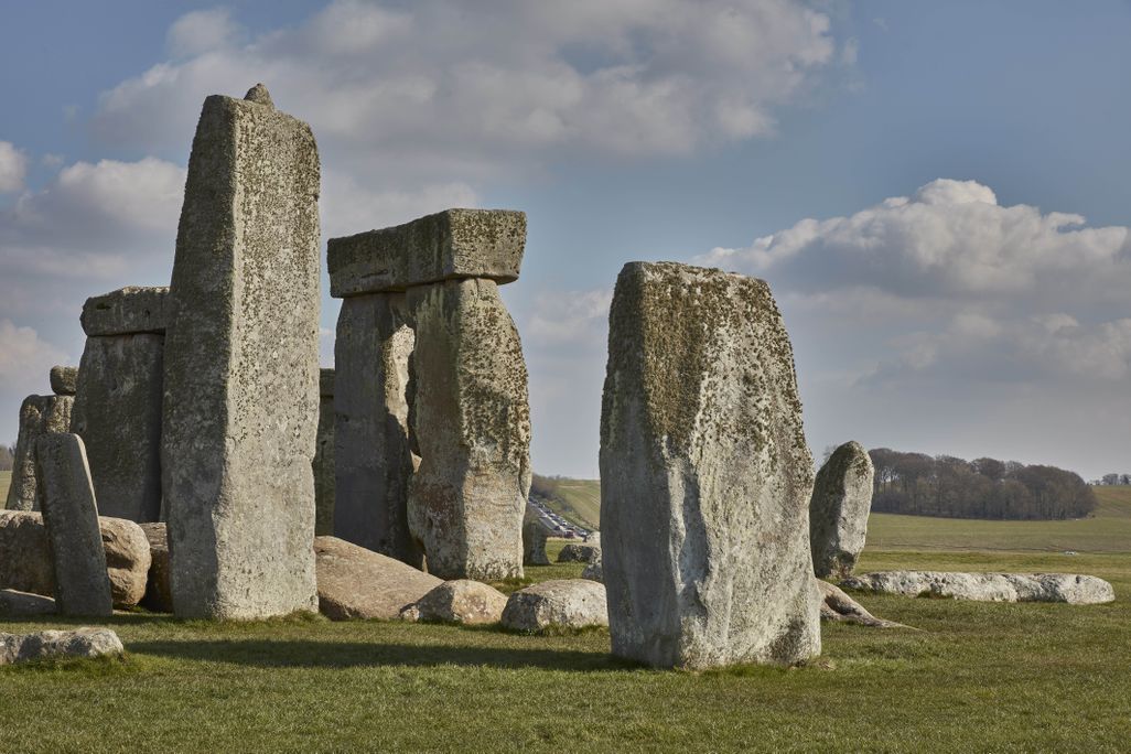 A view of Stonehenge's iconic square stones, under a blue sky with puffy white clouds; in the distance between two of the large stones, a trafficked road full of cars snakes over a hill