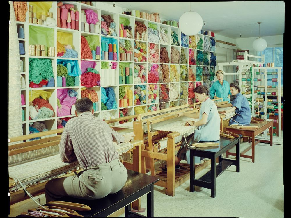 A smiling Dorothy Liebes stands in her studio in her blue apron, supervising two young women sitting at looms with spools of colorful thread and skeins of yarn close at hand.