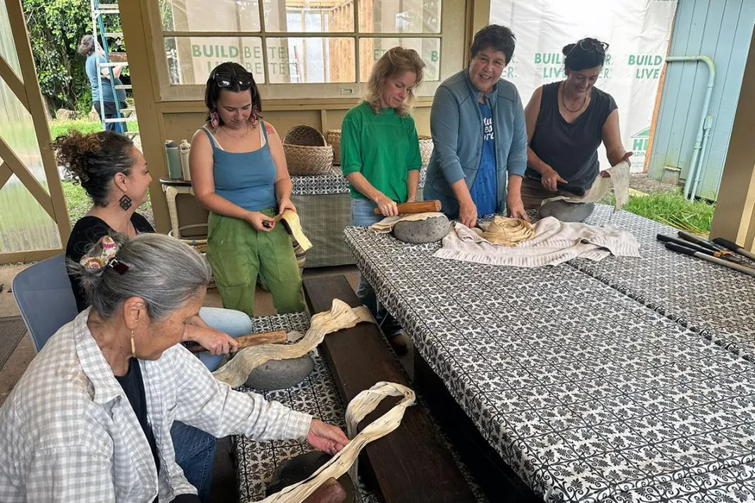 Six people sit and stand around a work table, each with a strip of pale bark cloth in their hands.