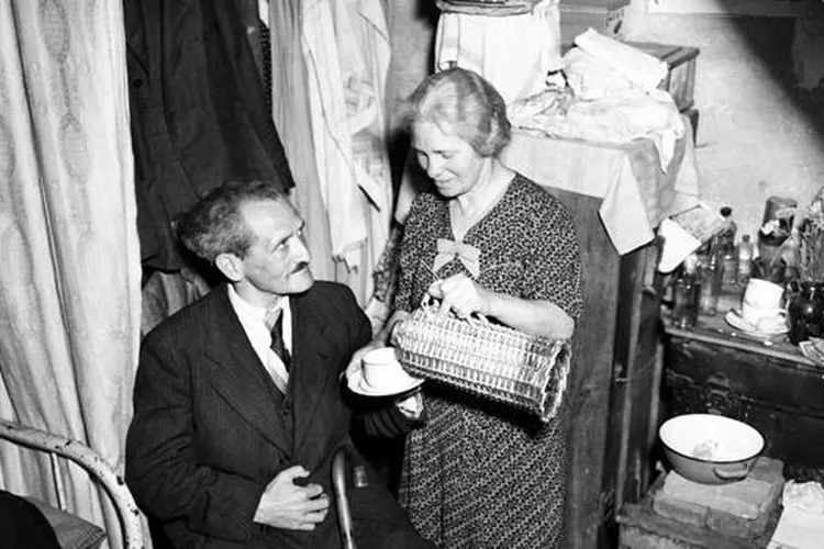 A white woman in a dress pours tea for a seated man in a suit, in a small home crowded with items (mobile)
