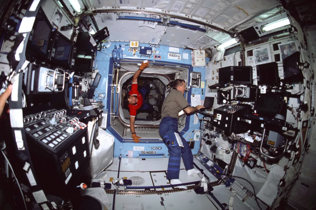 In 2001, astronaut James Voss performs a task at a work station in the International Space Station (ISS) Destiny Laboratory, as astronaut Scott Horowitz floats through the hatchway leading to the Unity node