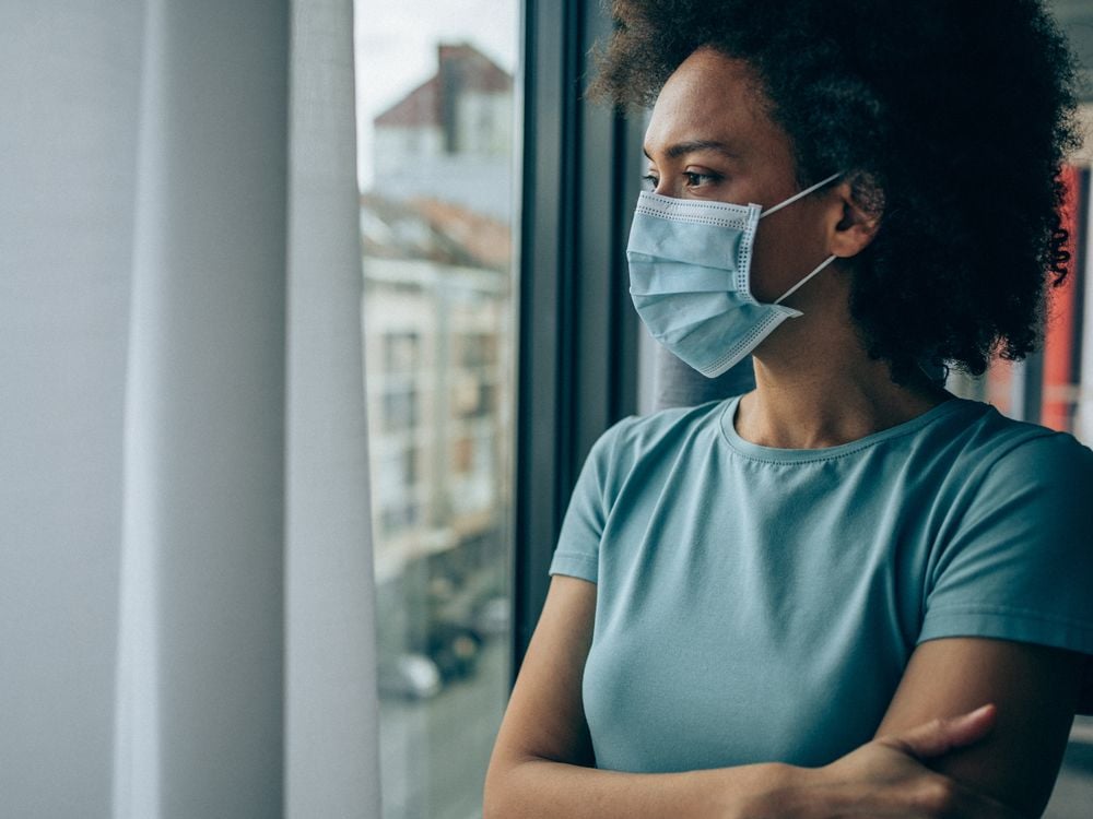 A Black woman with a face masks looks out of a window
