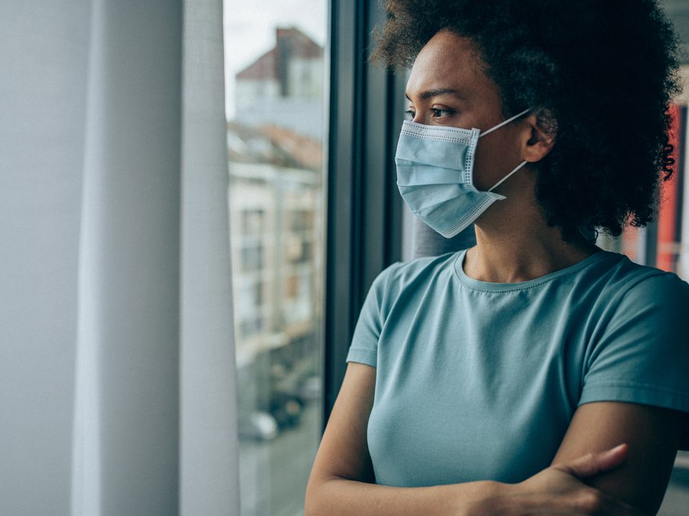 A Black woman with a face masks looks out of a window