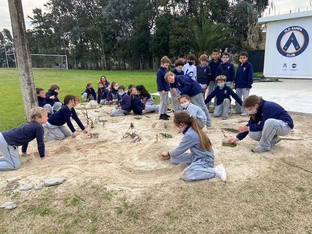 Students cluster in groups and individually around a sandy area, adding and refining the community map they are building out of sand, leaves, and sticks.