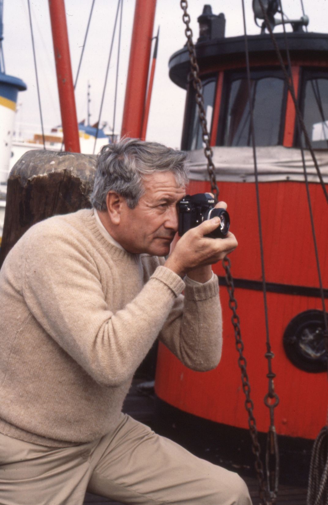 A man with grey hair and beige sweater holds a camera to his eye, presumably shooting photographs against a backdrop of marine craft.