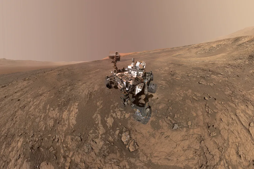 An image of the Mars Curiosity rover on planet Mars with Mt. Sharp in the background. On the right, towards the horizon is the Gale Crater's rim.