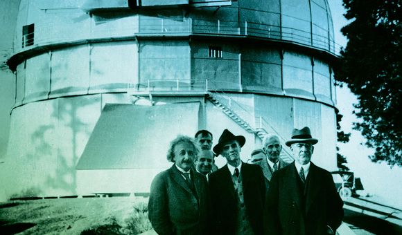 A black-and-white historical photograph from circa 1931 shows Albert Einstein with Edwin Hubble and other astronomers standing on a walkway in front of the large dome of the Mount Wilson Observatory.