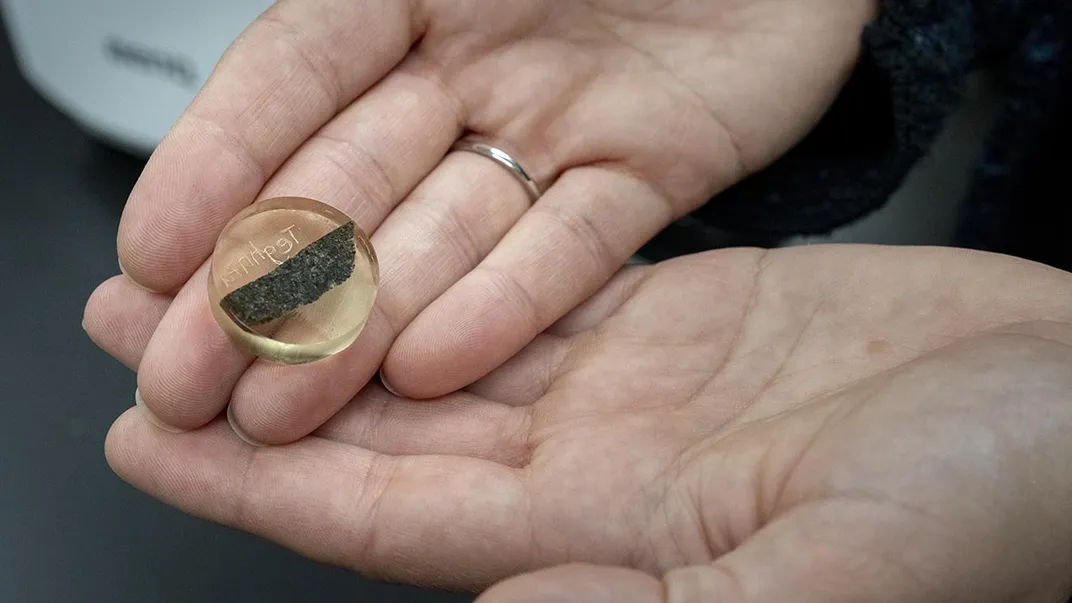 two hands holding a small piece of rock suspended in what looks like a glass cylinder