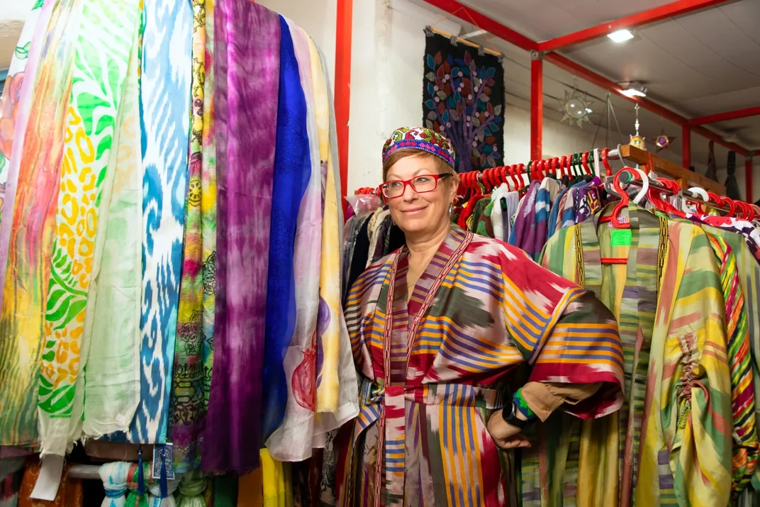 Lola Sayfi stands among clothing racks. She smiles to the side with her hands on her hips. The racks are filled with long, vibrant multicolored clothes.