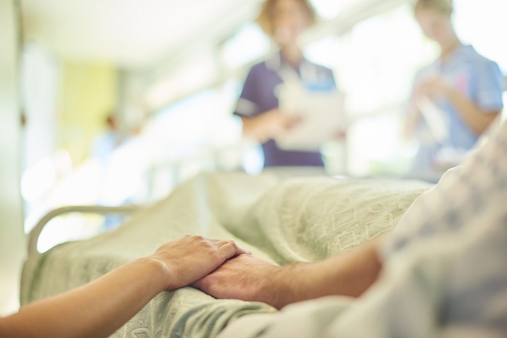 Close-up of person in hospital bed holding hands, with nurses blurred in the background