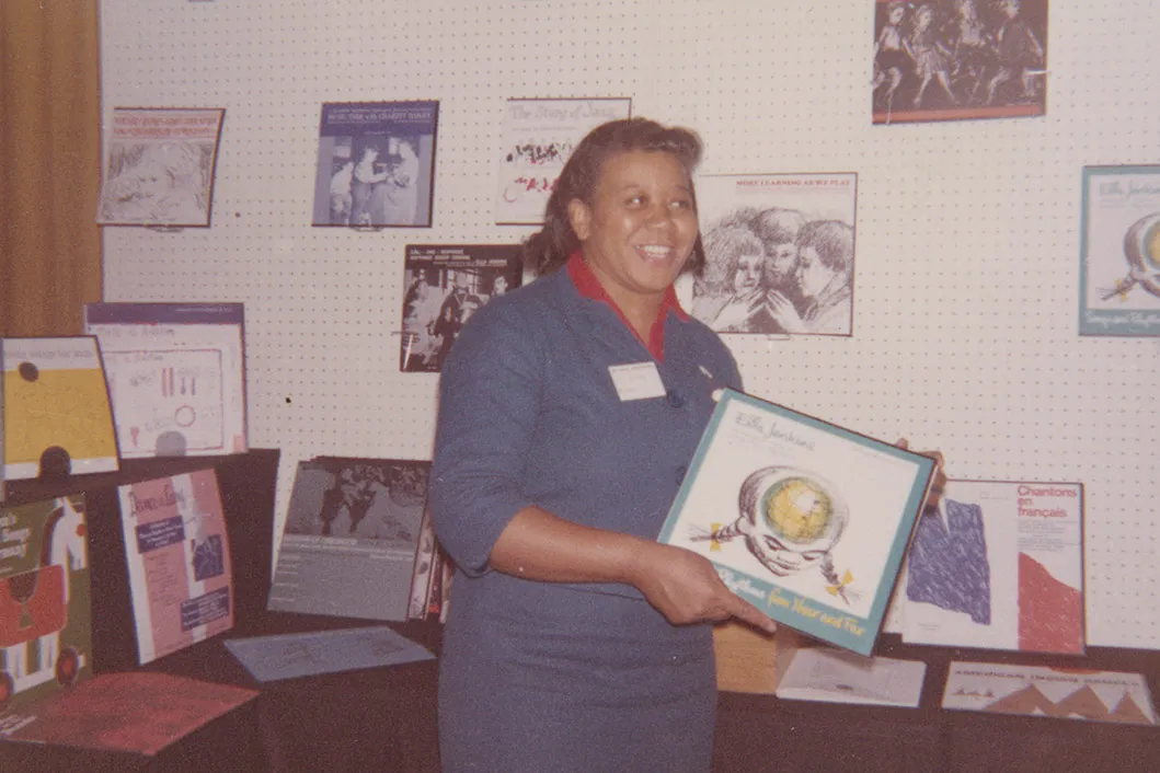 A woman with dark skin and long blue dress holds up a twelve-inch record. More records are on display behind her on a table and the wall.