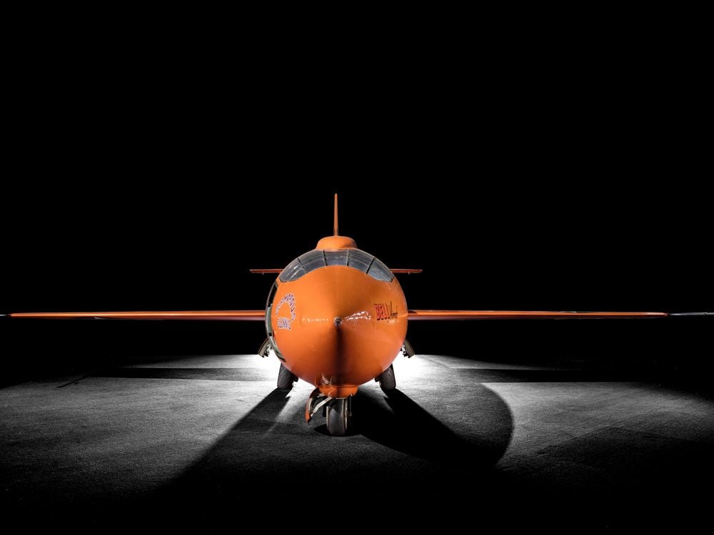 A bright orange plane, just big enough for one person to fit into, with cockpit facing the camera and each wing extended to either side, framed by dramatic lighting and a black background