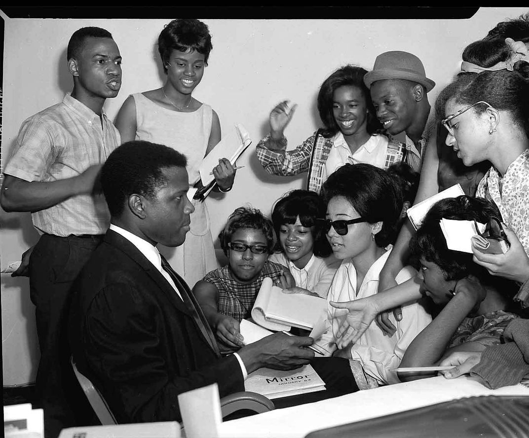Poitier wears a black suit, white shirt and dark tie and sits in front of an excited group of young Black people, who all appear to be clamoring for his autograph