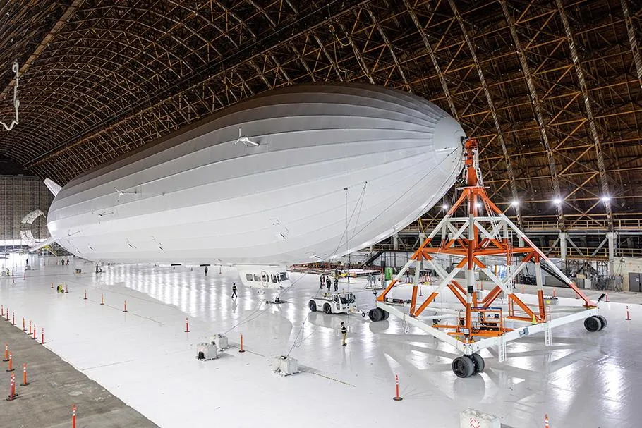 A large, white airship sits inside a hangar. People in safety jackets work on the ground.