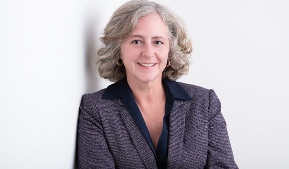 Elizabeth Babcock from the waist up against a white background. She is leaning against a wall with her arms crossed in front of herself.