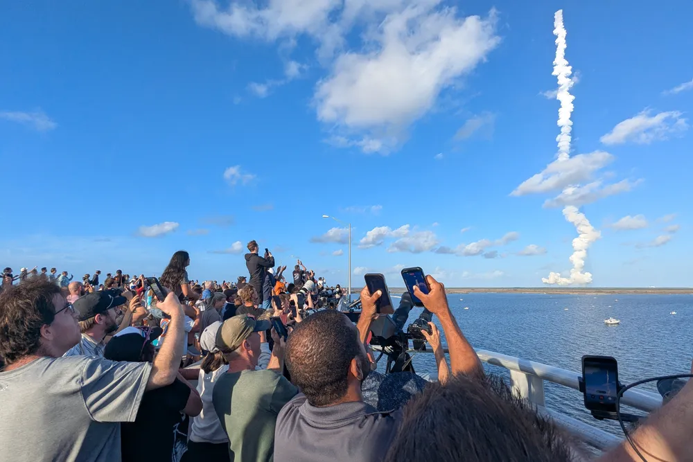 people raise their phones to take pictures of the rocket launch, seen in the distance as a plume of white steam