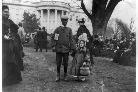 This 1898 photograph shows a young black boy holding hands with a young white girl during the Easter egg roll. The contraption on her head is an Easter bonnet. 