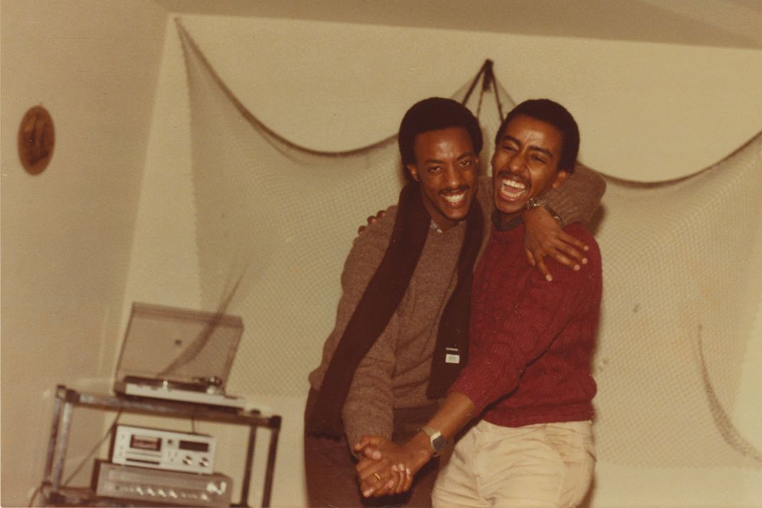 Two men embrace, as if dancing, while smiling and laughing, in what appears to be a basement room.