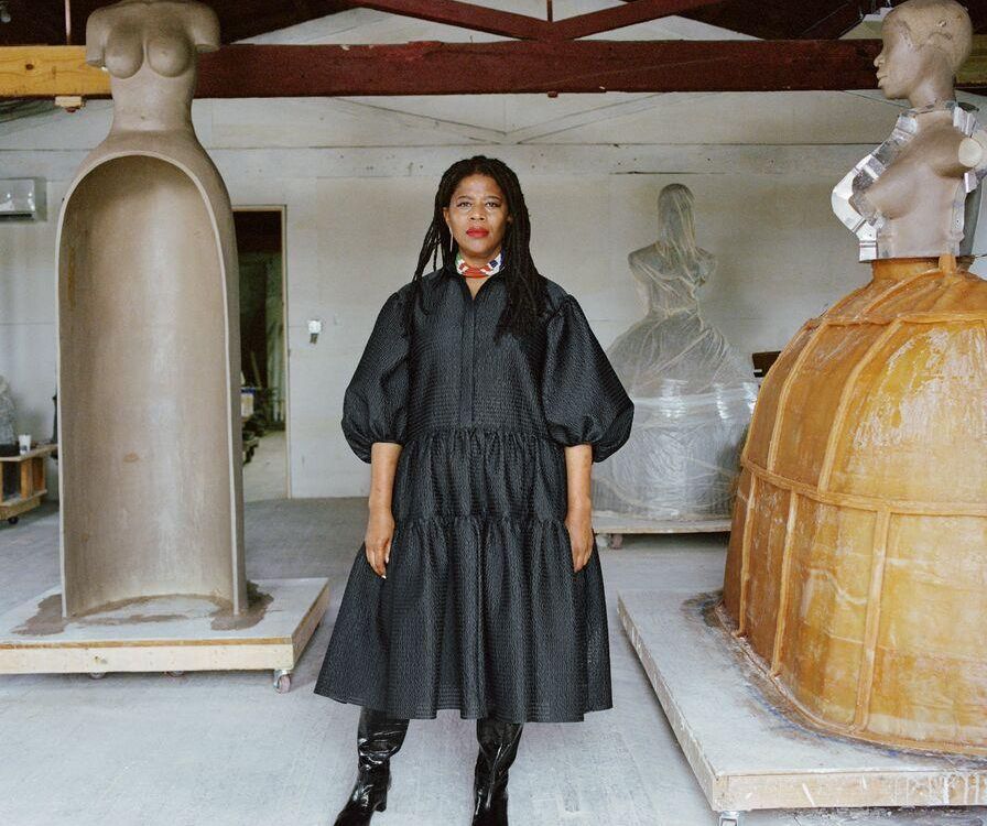 Simone Leigh, an African American woman, wears a voluminous black dress and stands in front of three of her sculptures, which are abstracted depictions of female figures on top of hoop skirts and jugs; Leigh faces the camera with a serious expression