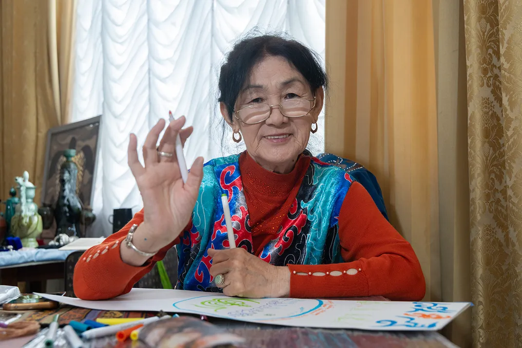 A woman wearing a red shirt and turquoise batik scarf smiles and waves, pencil in hand, while working on a drawing.